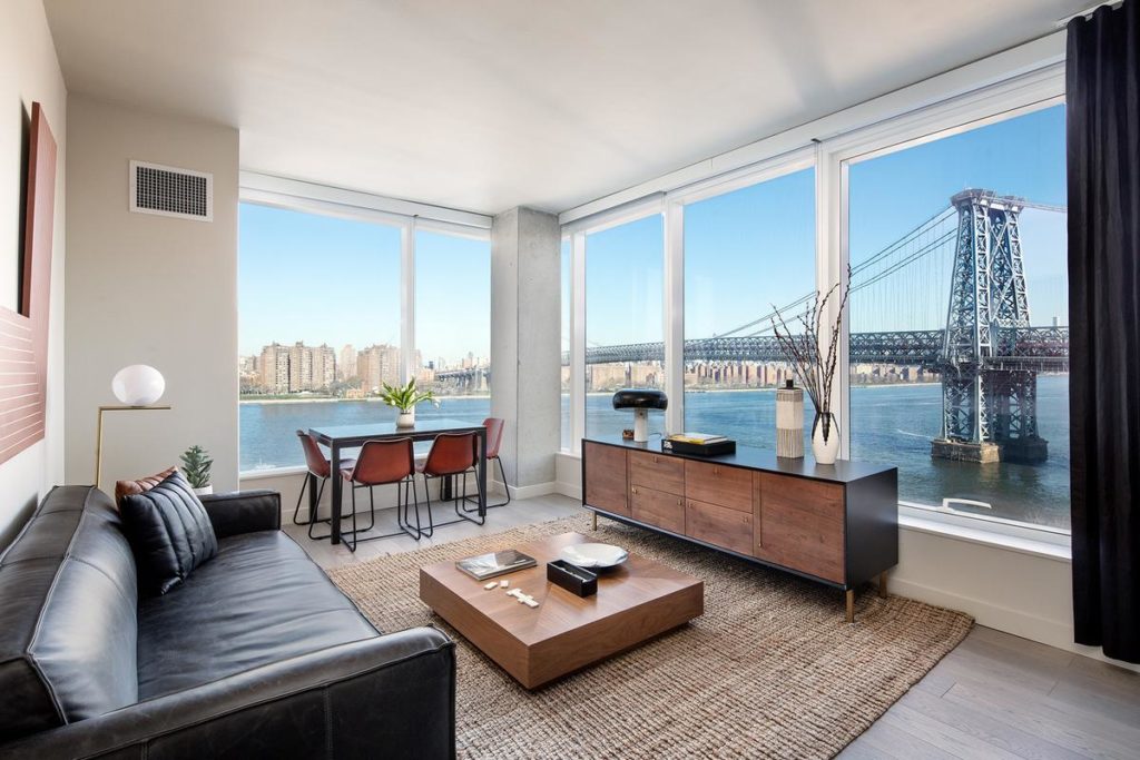 Bright Living Room with Couch, Wooden Media Stand and Coffee Table, Dining Table, Wood Flooring with Area Rug, surrounded by Oversized Windows with Williamsburg Bridge Waterfront and City Views